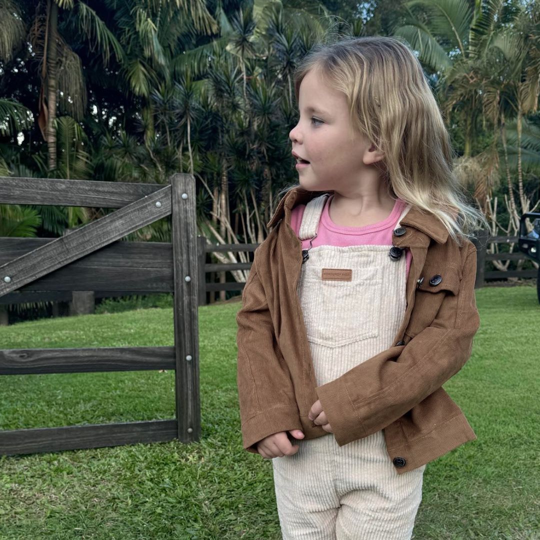 young girl wearing kids chocolate corduroy long sleeve dress shirt and beige corduroy overalls with a back drop of beautiful grass and a fence line